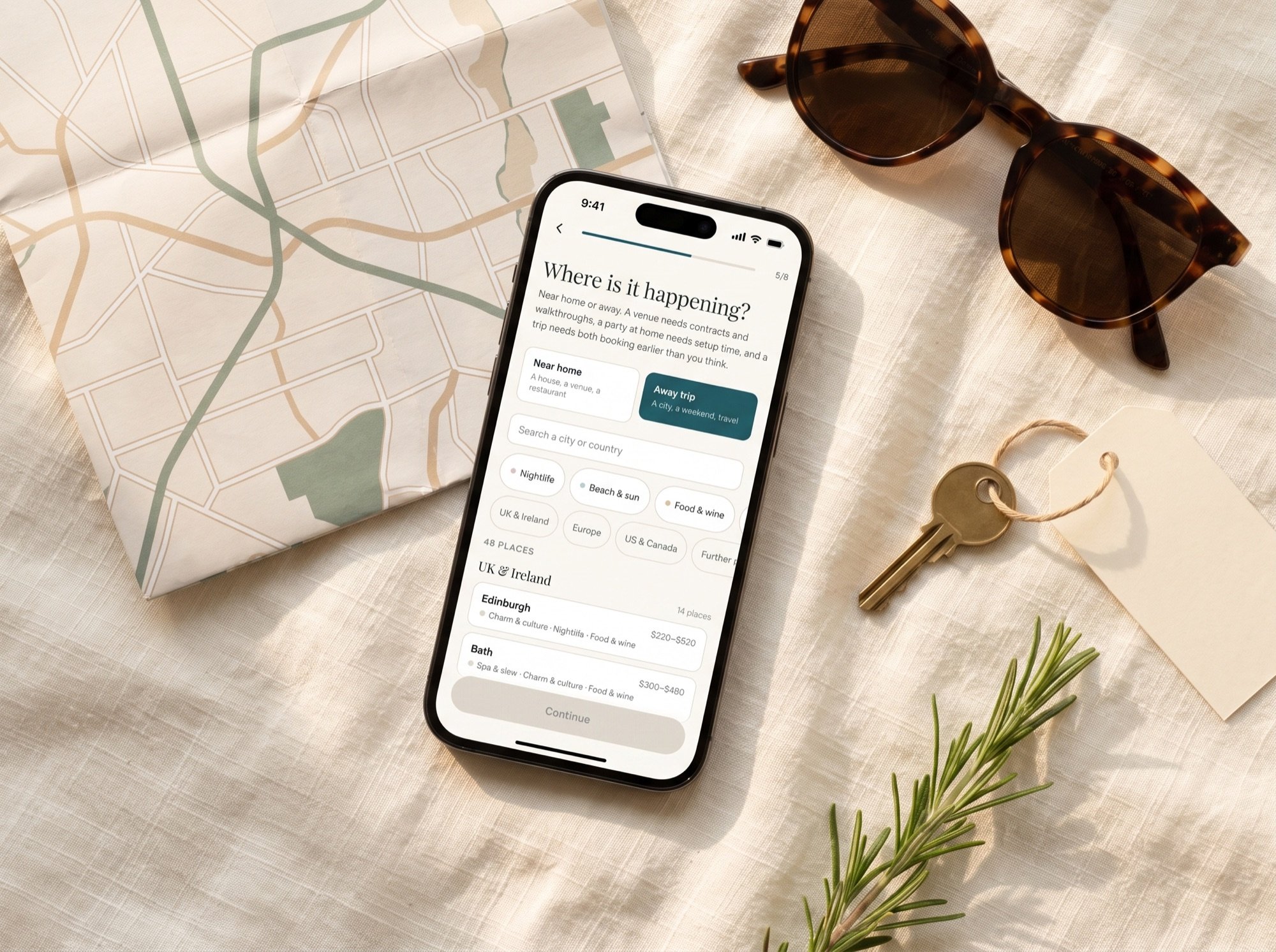 A phone showing the planner's where-is-it-happening step, on cream linen with a folded map, sunglasses, a key on a blank tag and a sprig of rosemary